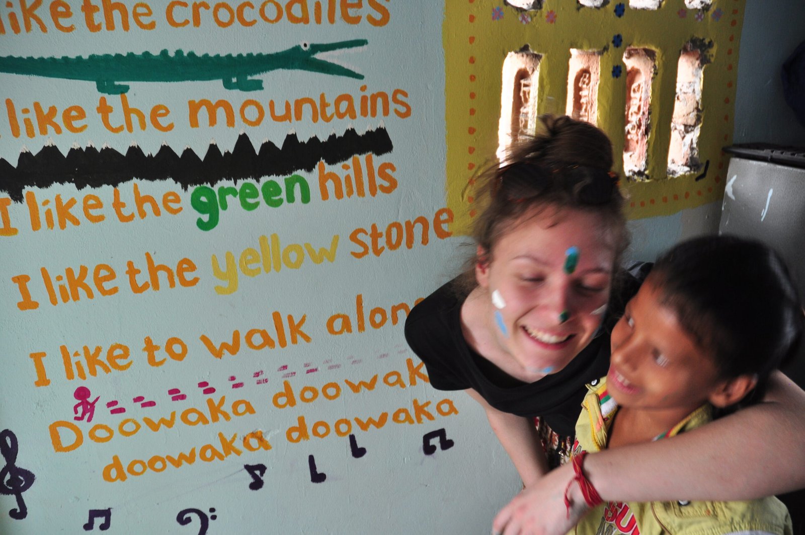 A volunteer with festival colors on her face sharing a laugh with a boy beside a painted classroom wall
