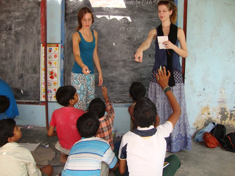 Two volunteers teaching at a blackboard while children sitting on the floor raise their hands