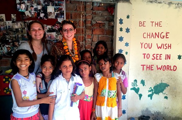 Two volunteers with girls beside a wall painted with “Be the change you wish to see in the world”